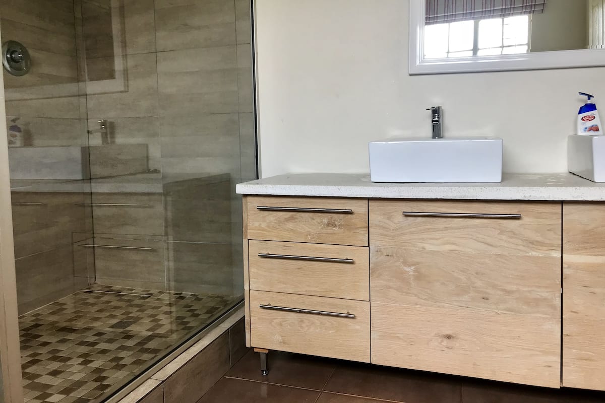 A bathroom with a glass shower enclosure, a white sink, and wooden cabinets.