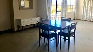 A dining area with a table and chairs, a white cabinet, and a window with curtains. - Windmill Farm (Jagersrust)