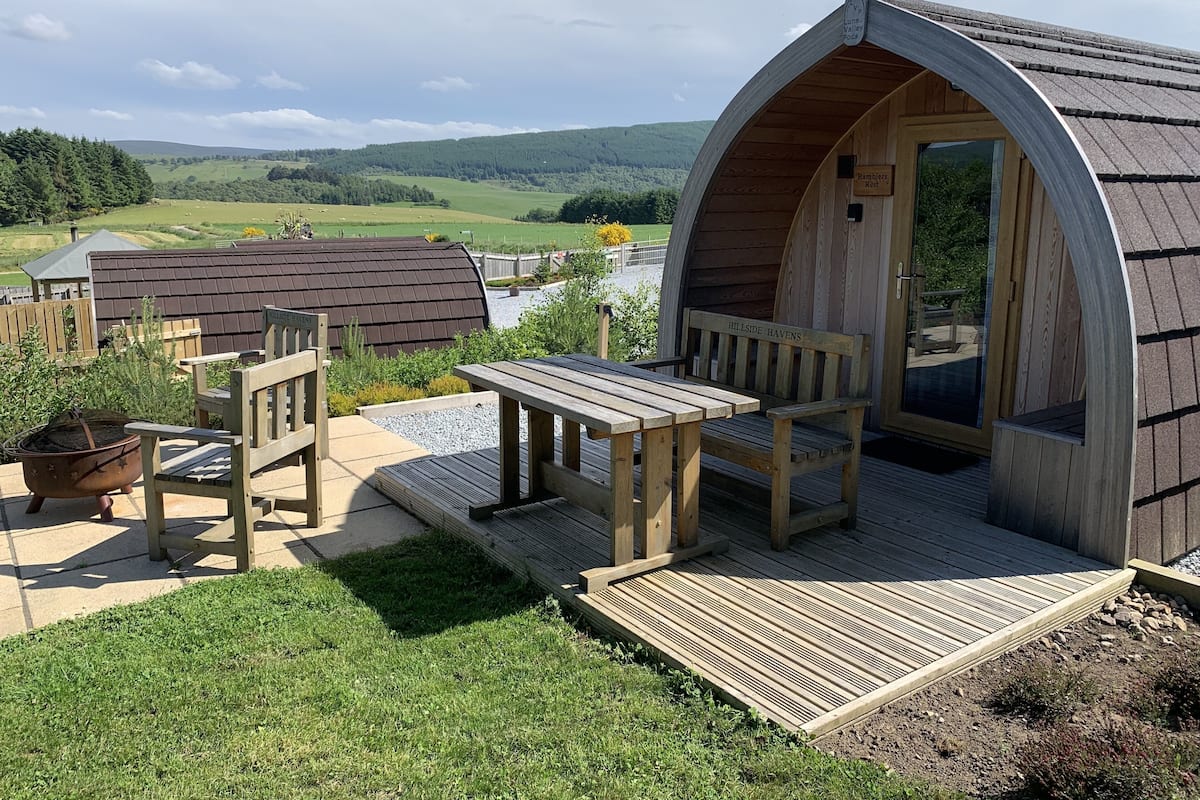 A wooden cabin with a curved roof, a bench, a table, and a fire pit.