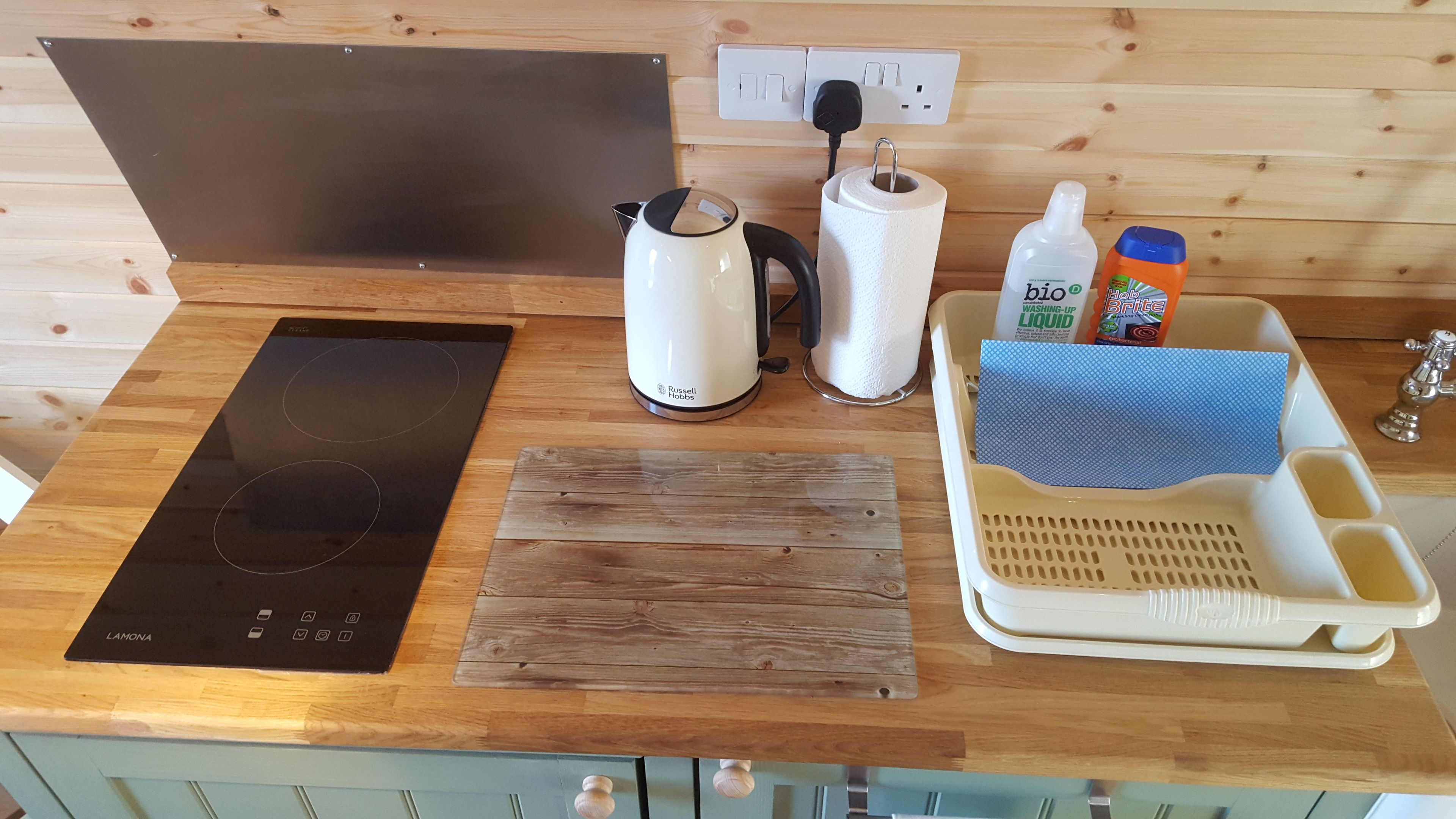 A kitchen counter with a black induction cooktop, a white kettle, a roll of paper towels, a bottle of dish soap, and a blue cloth in a tray.