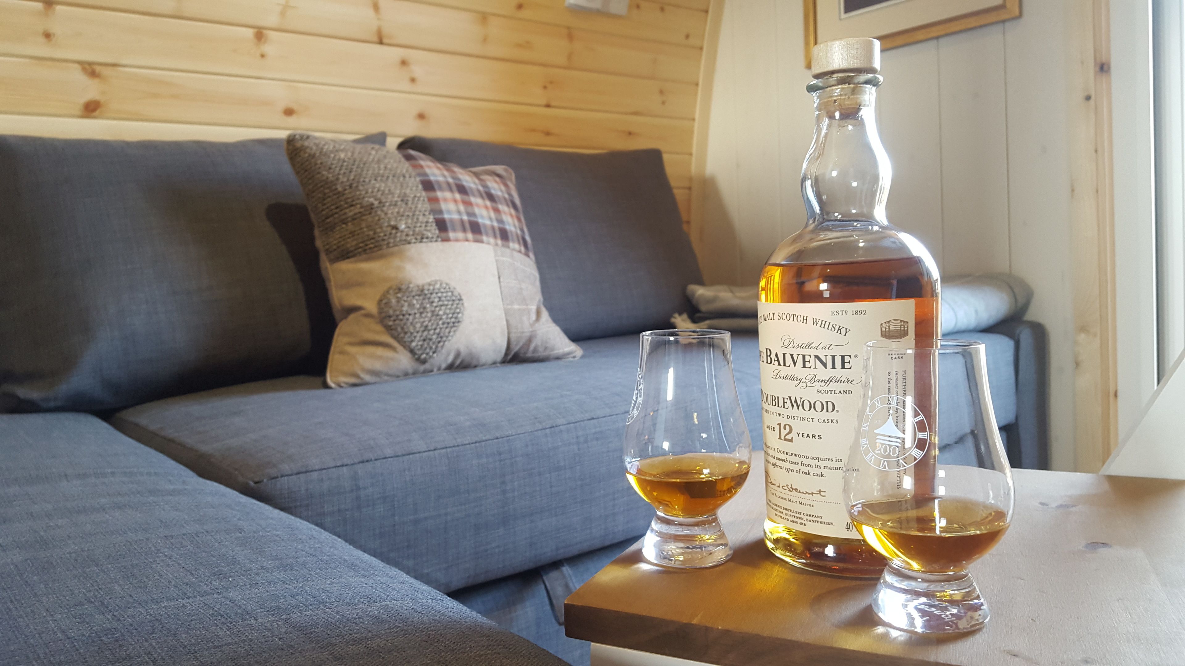 A bottle of Doublewood Malt Scotch Whisky and two glasses on a wooden table in a room with a blue sofa and a plaid pillow.