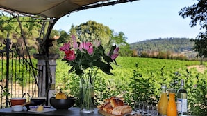 A table set outdoors with a view of a vineyard, a vase of flowers, and various food and drink items. - La Belugue (Beaumont-de-Pertuis)