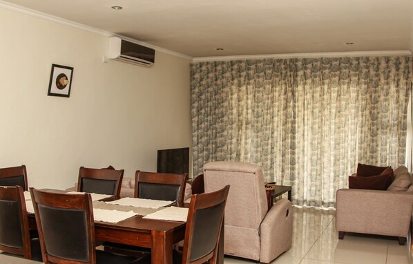 A dining area with a wooden table and chairs, a sofa, and a large window with curtains. - Apartments at 14 Mombo Road (Blantyre)