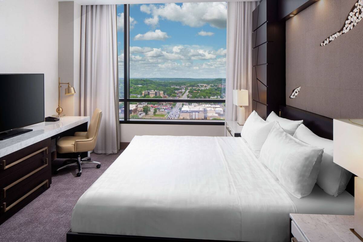 A hotel room with a large bed, a desk with a chair, a TV, and a view of a cityscape through a large window.