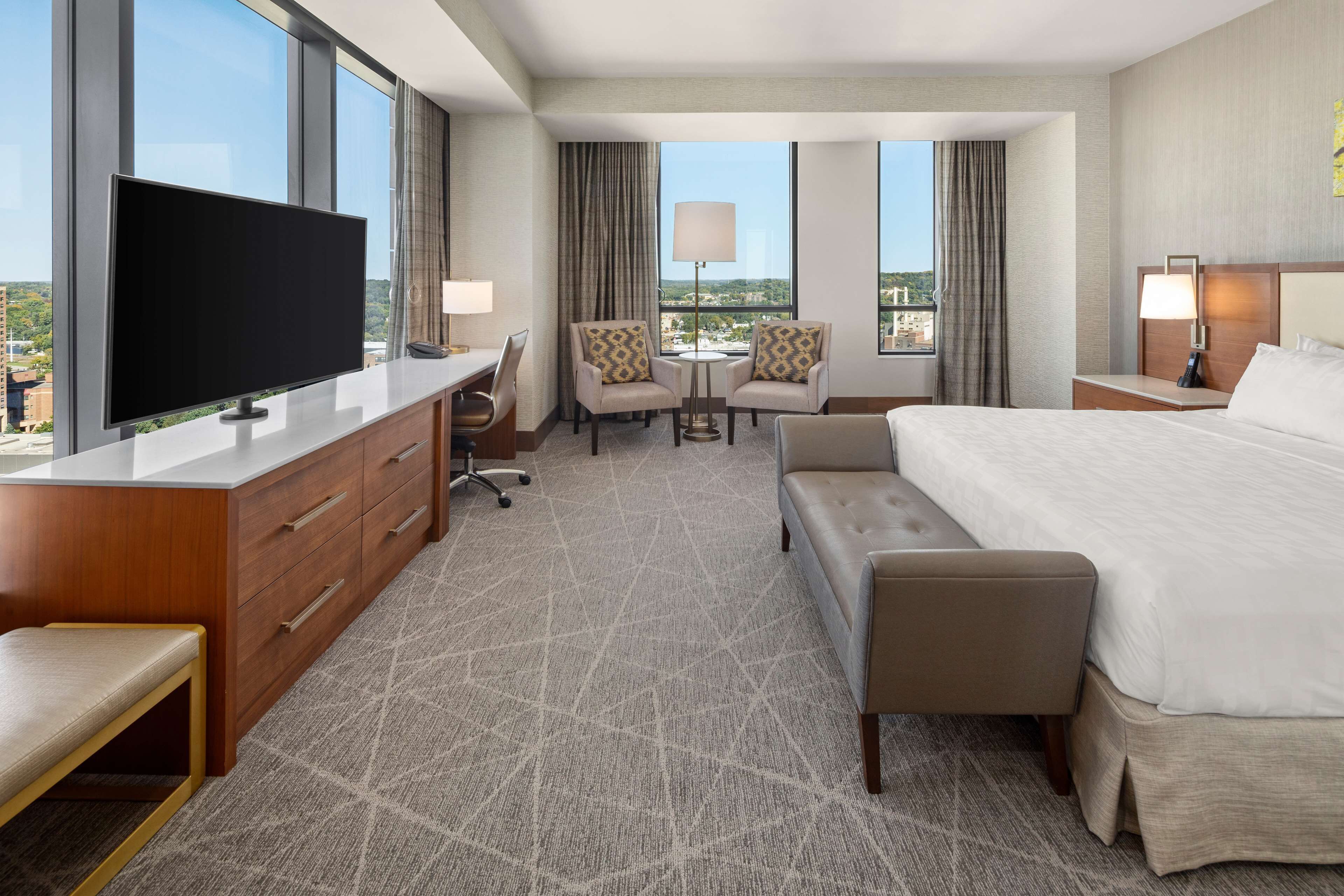 A hotel room with a large flat-screen TV, a desk with drawers, two armchairs, a round table, a bed with a headboard, and a view of the cityscape through large windows.