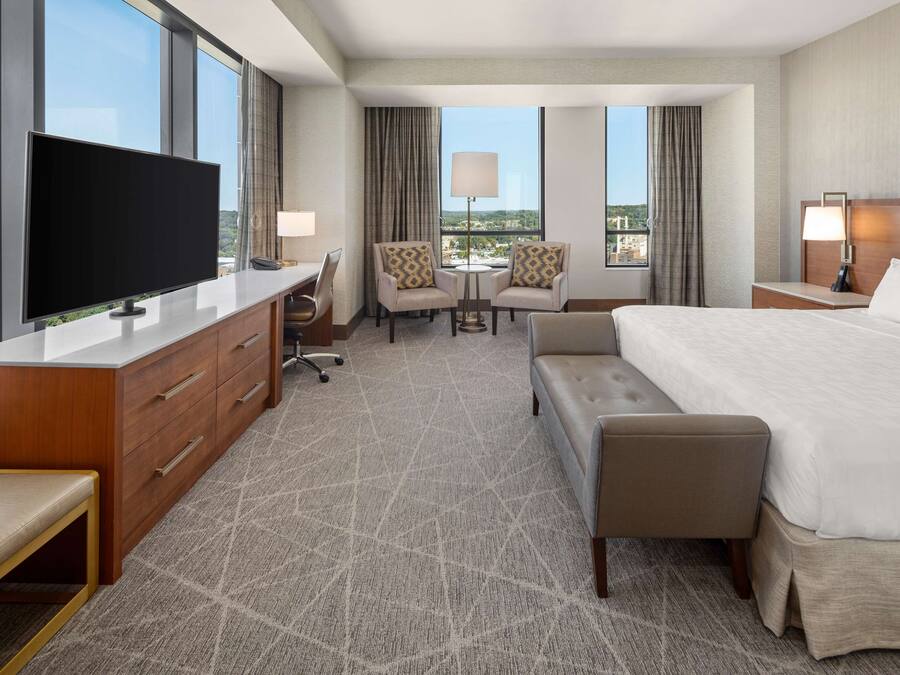 A hotel room with a large flat-screen TV, a desk with drawers, two armchairs, a round table, a bed with a headboard, and a view of the cityscape through large windows.