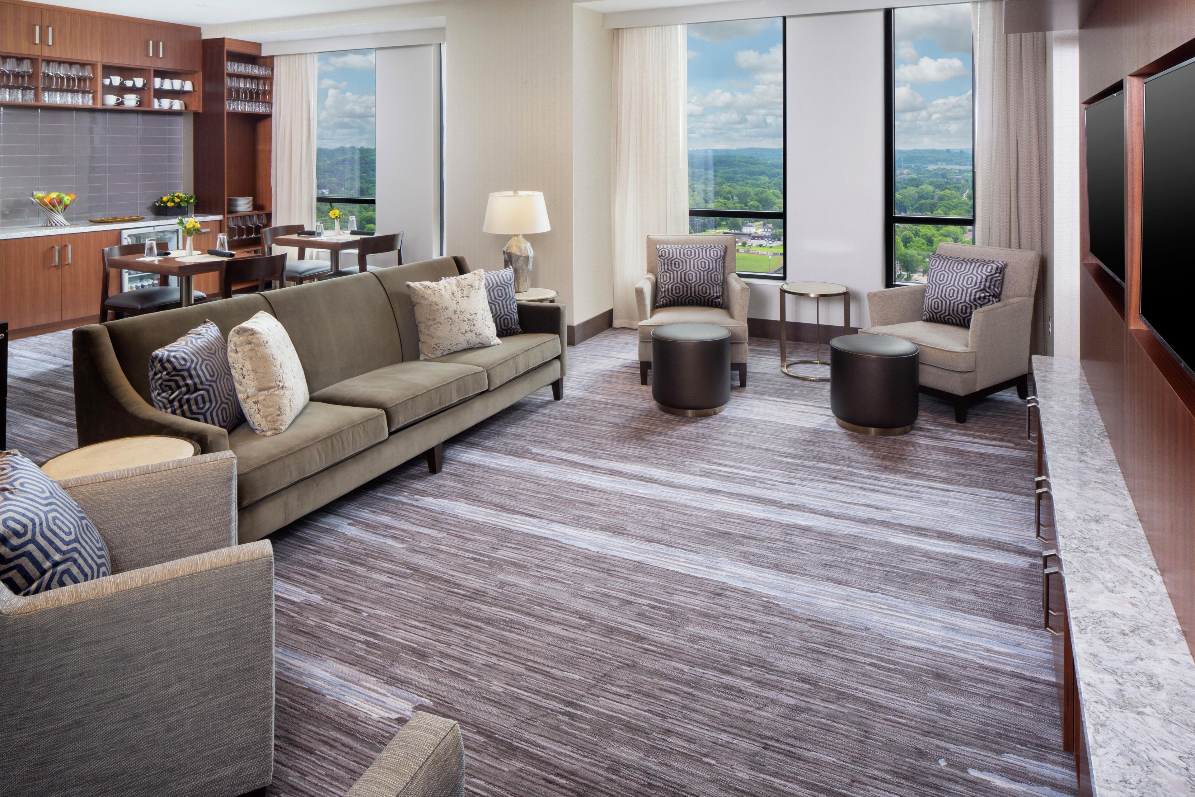 A modern hotel room with a living area, dining space, and a large window offering a city view.