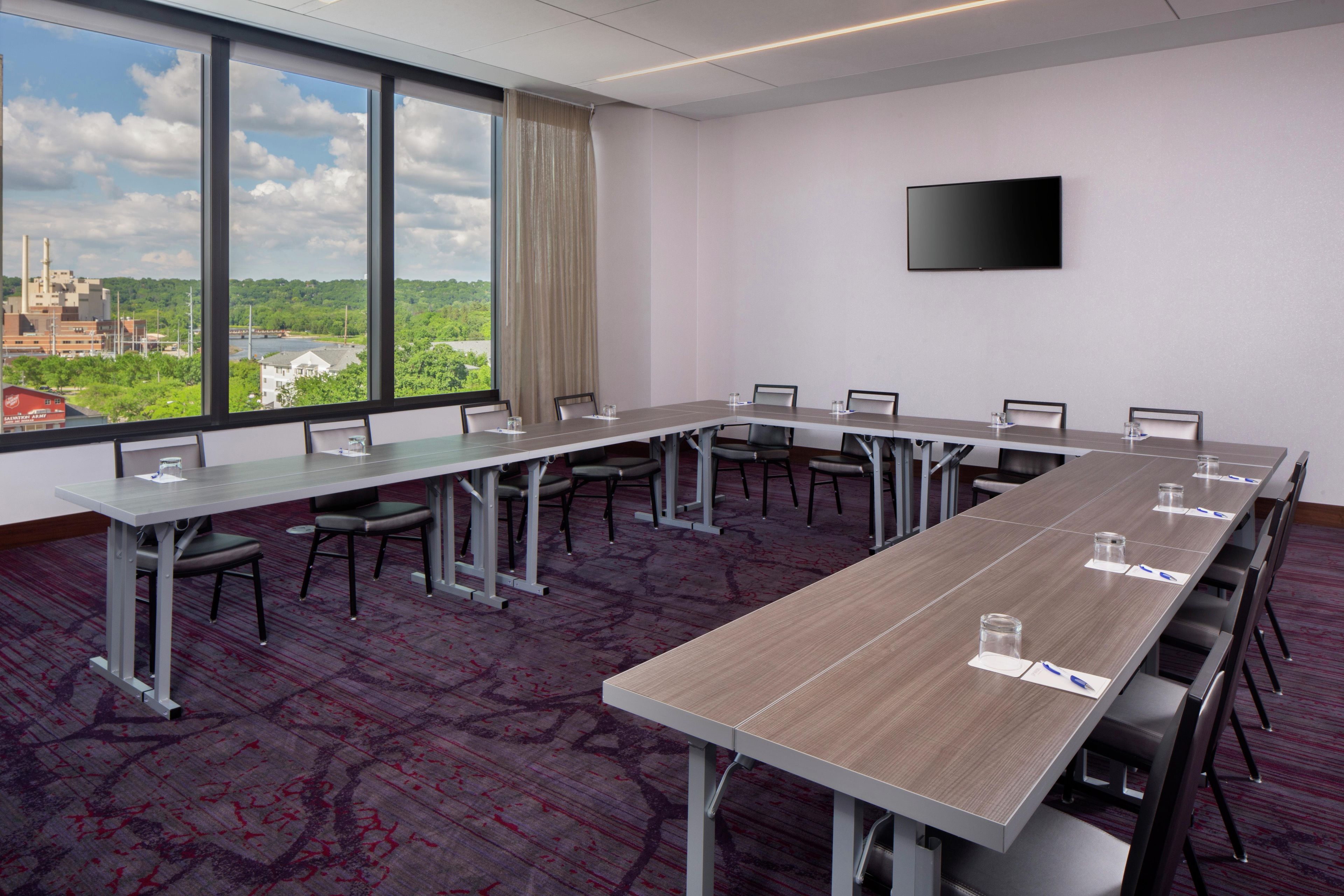 Un salón de conferencias con una larga mesa, sillas y un amplio ventanal con vistas a un edificio y vegetación.