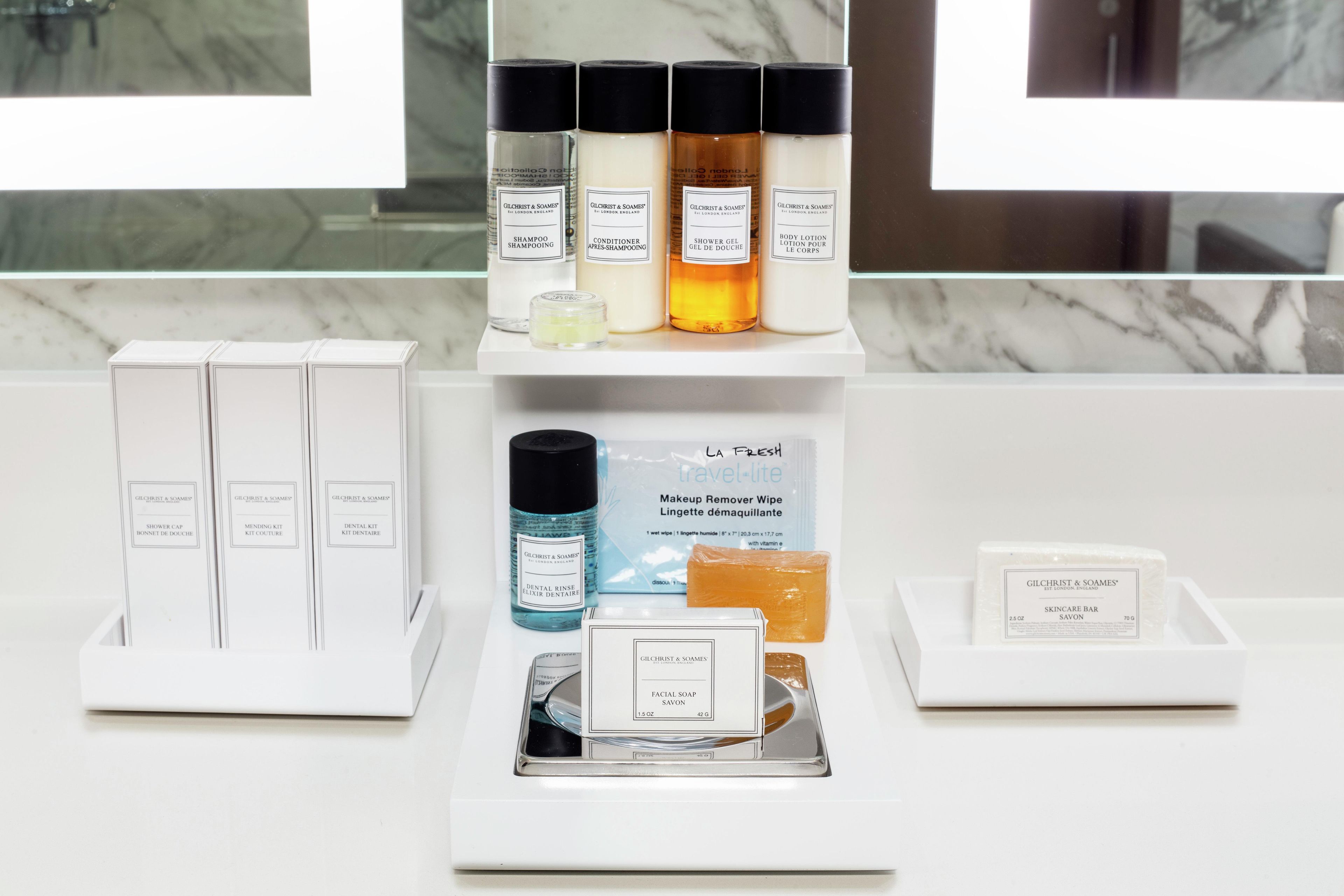 A bathroom vanity with toiletries including shampoo, conditioner, shower gel, body lotion, dental rinse, makeup remover wipe, facial soap, and skincare bar.
