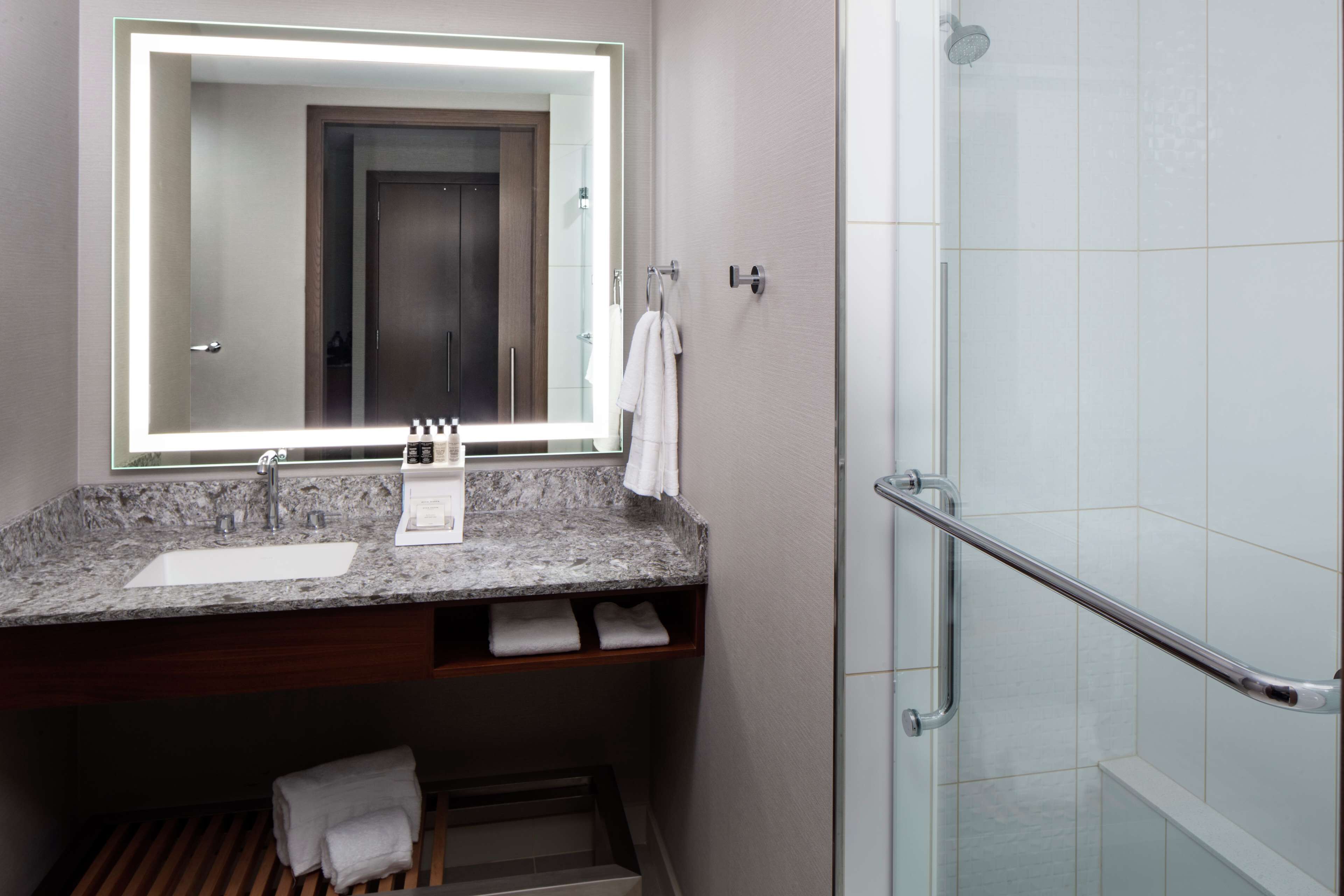 A bathroom with a granite countertop sink, a mirror with a light, a shower with a glass door, and a towel rack.