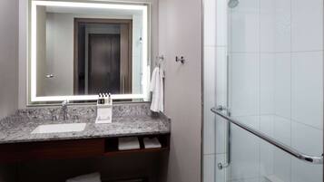 A bathroom with a granite countertop sink, a mirror with a light, a shower with a glass door, and a towel rack.