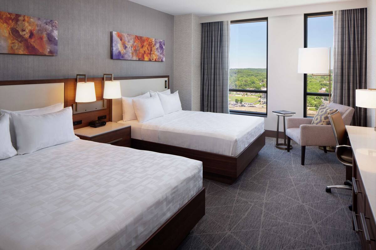 A hotel room with two beds, a desk, a chair, and a view of the outdoors.