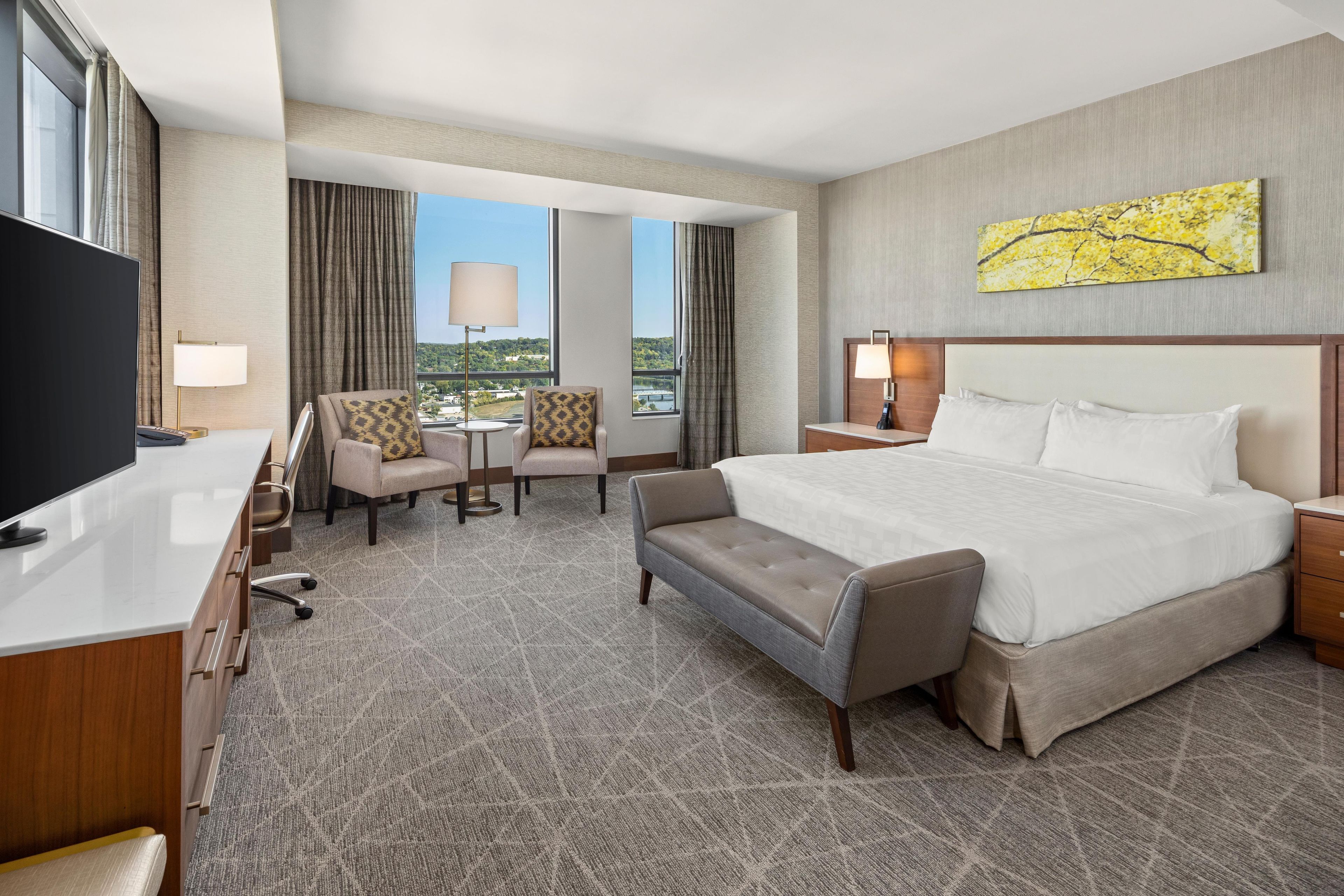 A hotel room with a large bed, a desk, a chair, a small table, and a TV.