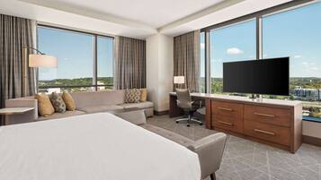A hotel room with a large bed, a sofa, a desk with a chair, a television, and large windows offering a city view.