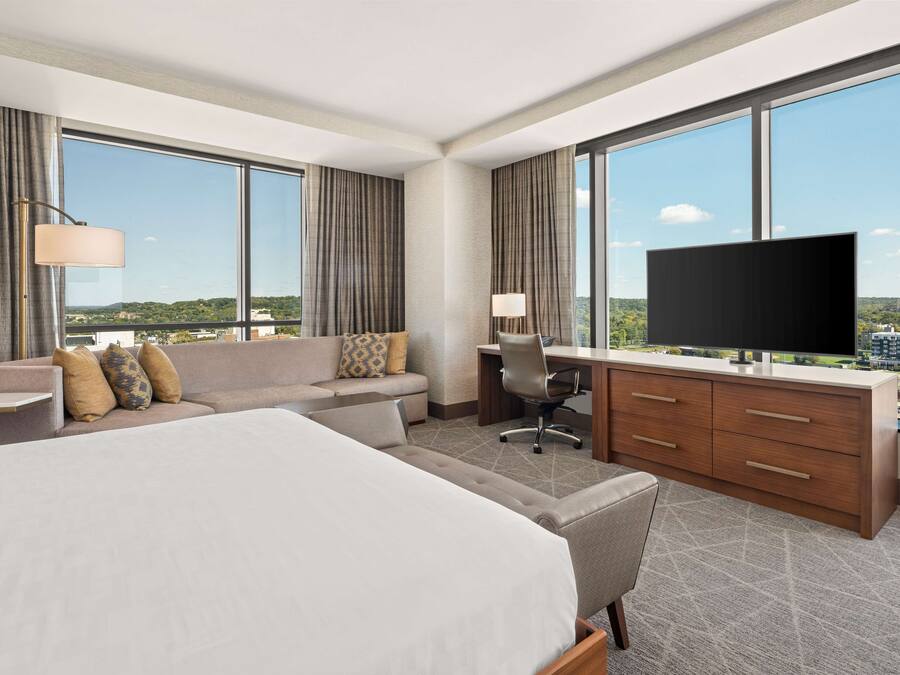 A hotel room with a large bed, a sofa, a desk with a chair, a television, and large windows offering a city view.
