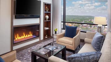 A modern living room with a flat-screen TV, a fireplace, a sofa, a coffee table, and a view of a cityscape through large windows.