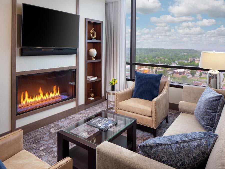 A modern living room with a flat-screen TV, a fireplace, a sofa, a coffee table, and a view of a cityscape through large windows.