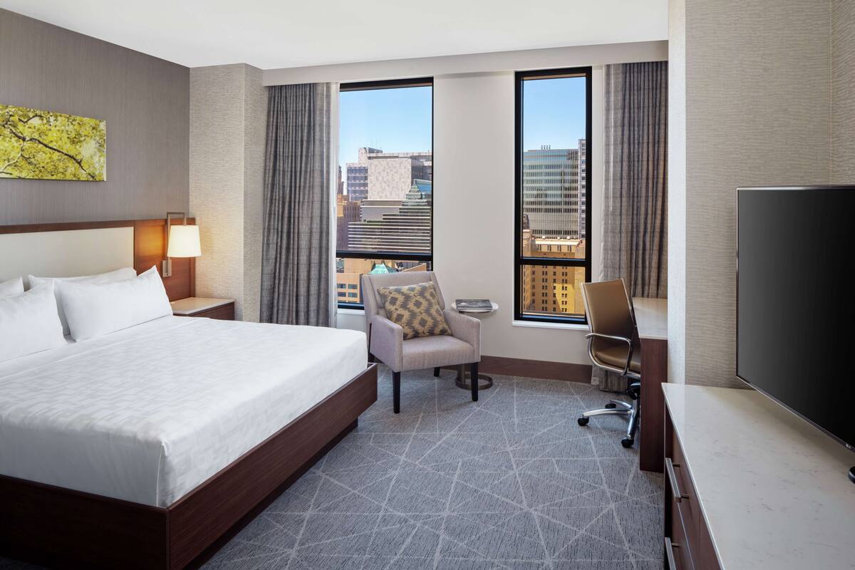 A hotel room with a large bed, a desk with a chair, a television, and a view of the city through large windows.