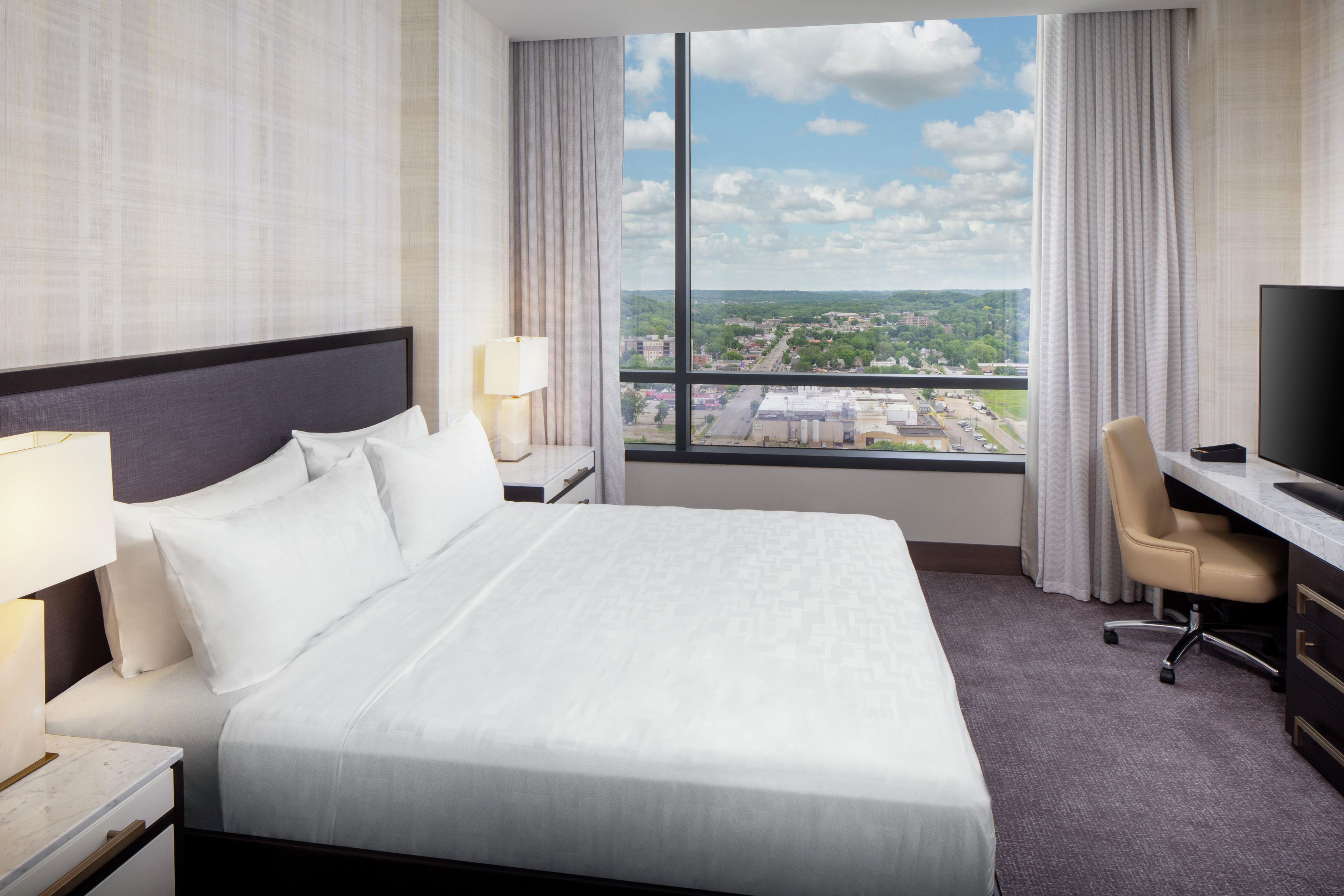 A hotel room with a large bed, a desk with a chair, a television, and a window with a city view.