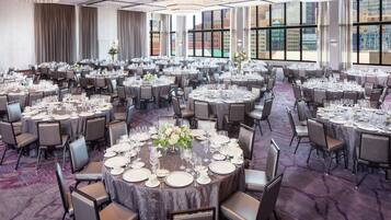 A large banquet hall with round tables set for dining, featuring a carpeted floor and large windows offering city views.