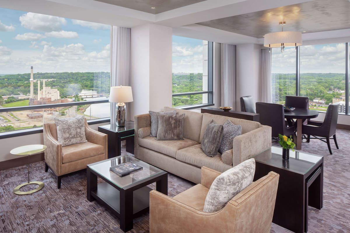 A modern hotel room with a large window offering a city view, a sofa set, a dining area, and a small side table.
