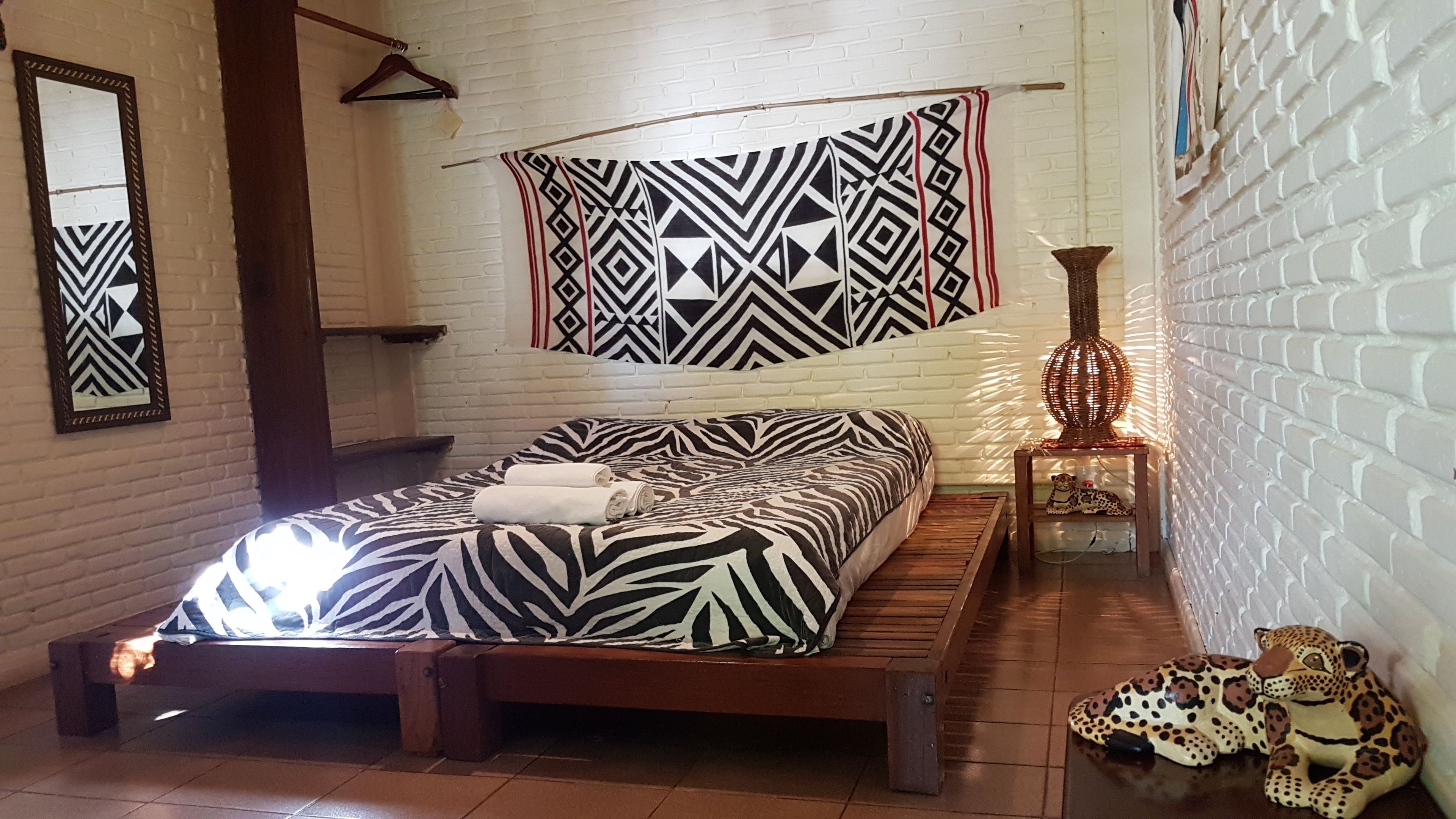 A room with a bed, a hanging tapestry, a small table with a lamp, and a decorative item on the floor.
