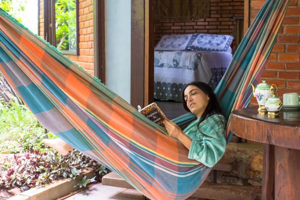 Uma pessoa relaxando em uma rede, lendo um livro, com um muro de tijolos e uma cama ao fundo.