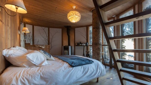 A cozy wooden cabin bedroom with a bed, a ladder, and a large window overlooking trees.