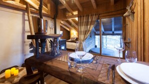 A wooden dining table set with wine glasses and plates, a chandelier, and a rustic interior with wooden beams and a view through a window.