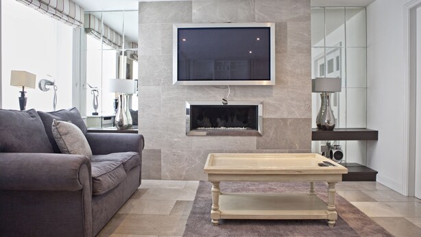 Una sala de estar moderna con un sofá gris, una mesa de centro de madera, un televisor de pantalla plana montado en una pared de mármol y una chimenea debajo del televisor.