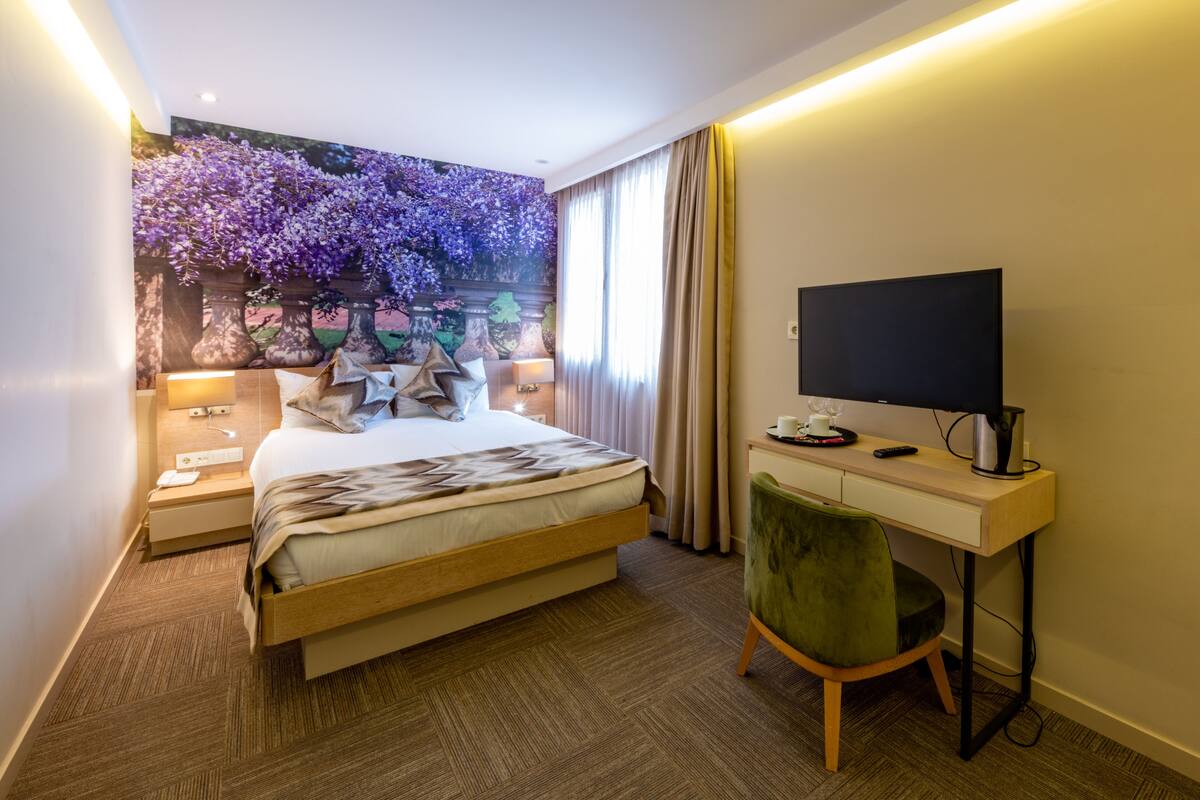 A hotel room with a bed, a desk with a computer, a TV, and a large floral mural on the wall.