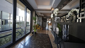 Un hall d’hôtel moderne avec un sol en marbre, de grandes fenêtres et un comptoir d’accueil agrémenté de plantes décoratives.
