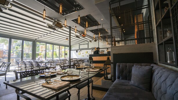 Un vaste espace dédié à la salle à manger, avec de longues tables, un éclairage moderne suspendu et un coin salon confortable agrémenté d’un banc rembourré.