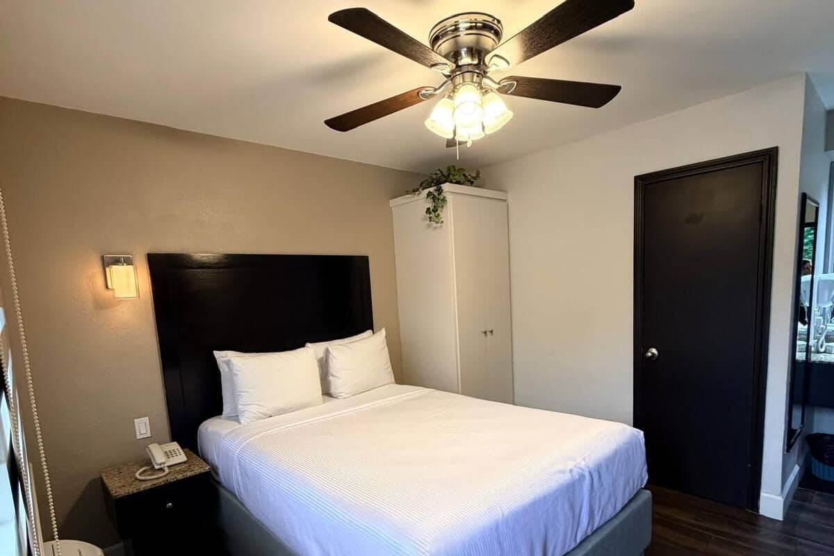 Une chambre d’hôtel comprenant un lit, un ventilateur de plafond, une table de chevet avec un téléphone, un radiateur fixé au mur et une armoire.