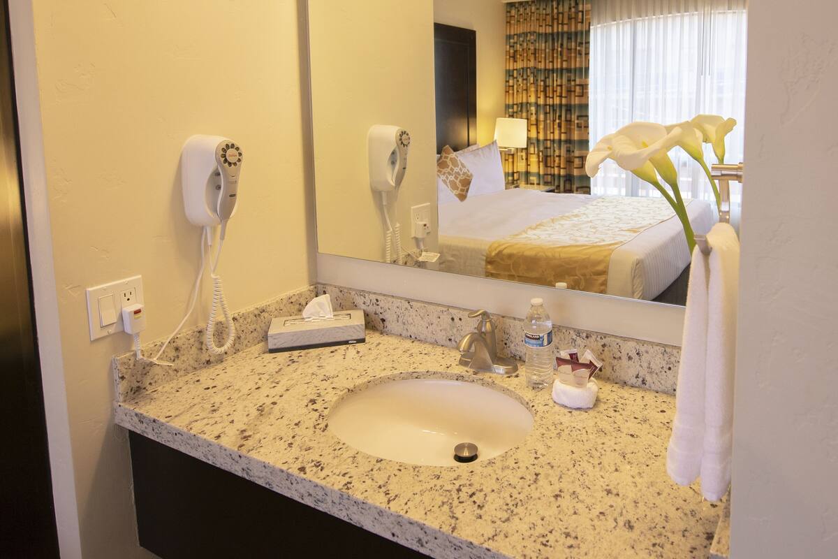 Une salle de bain d’hôtel avec un comptoir en granit, un lavabo, un miroir, un sèche-cheveux, une serviette et un lit visible dans le reflet.