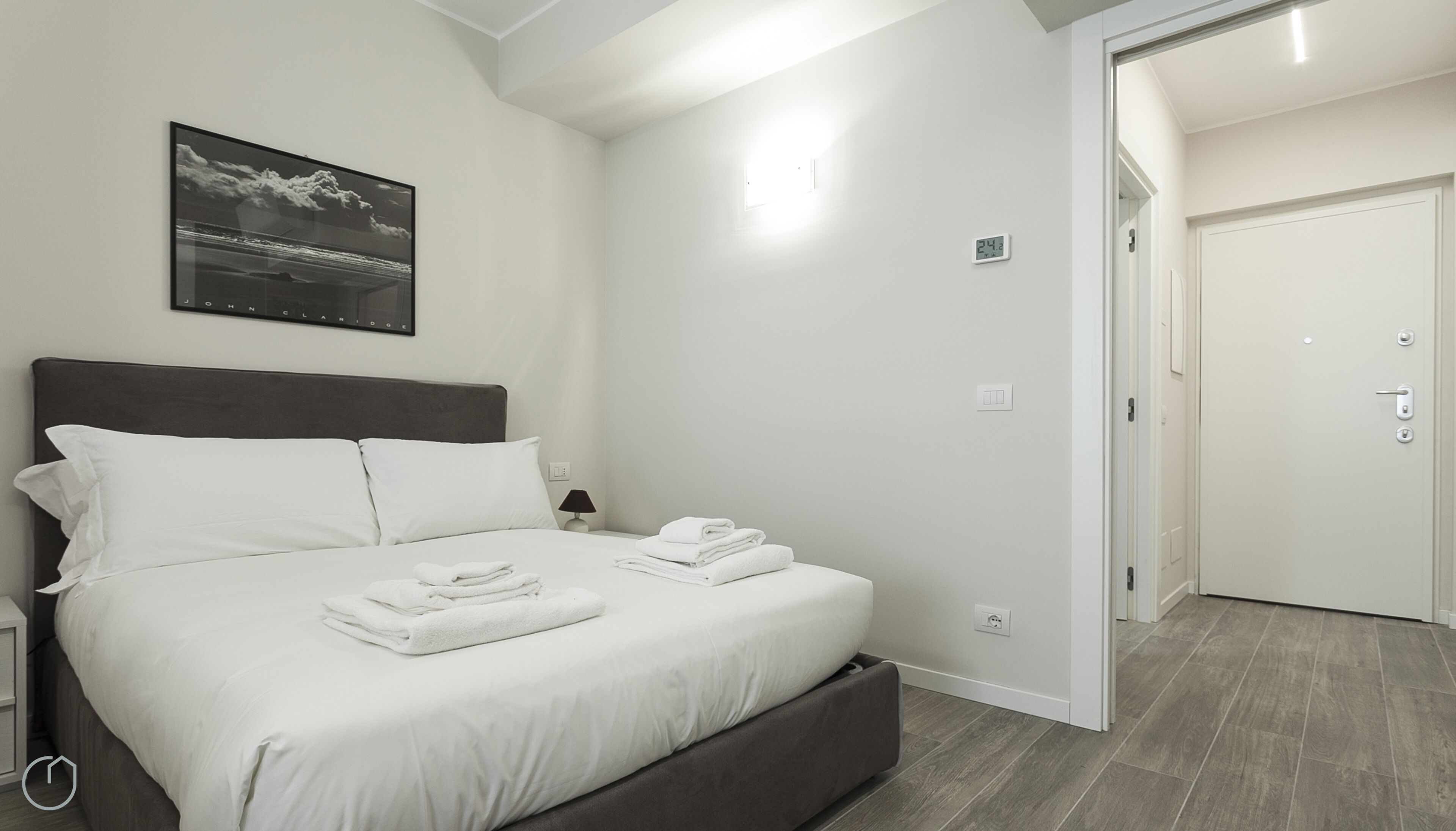 A modern bedroom with a large bed, a nightstand, a wall-mounted clock, and a door leading to another room.