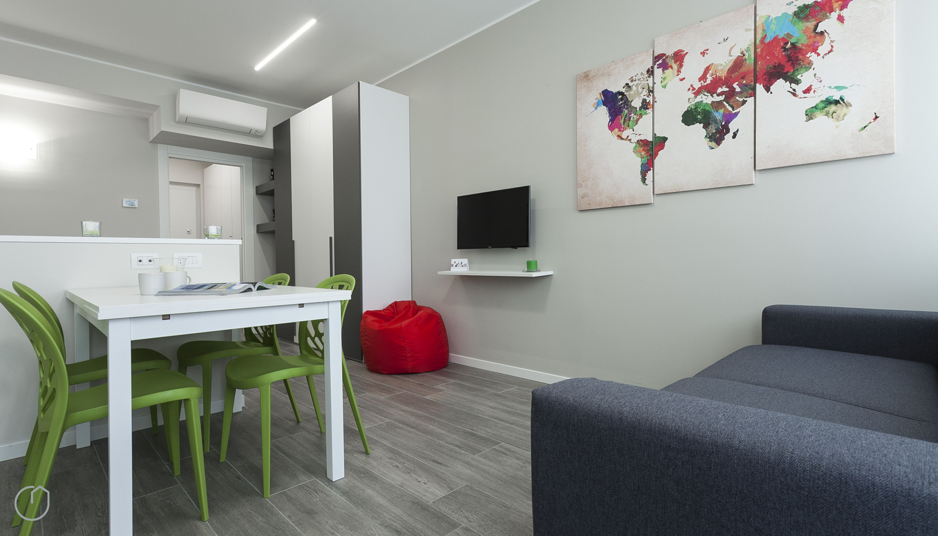 A modern, well-lit room with a dining area, a red bean bag, a grey sofa, and a flat-screen TV mounted on the wall.