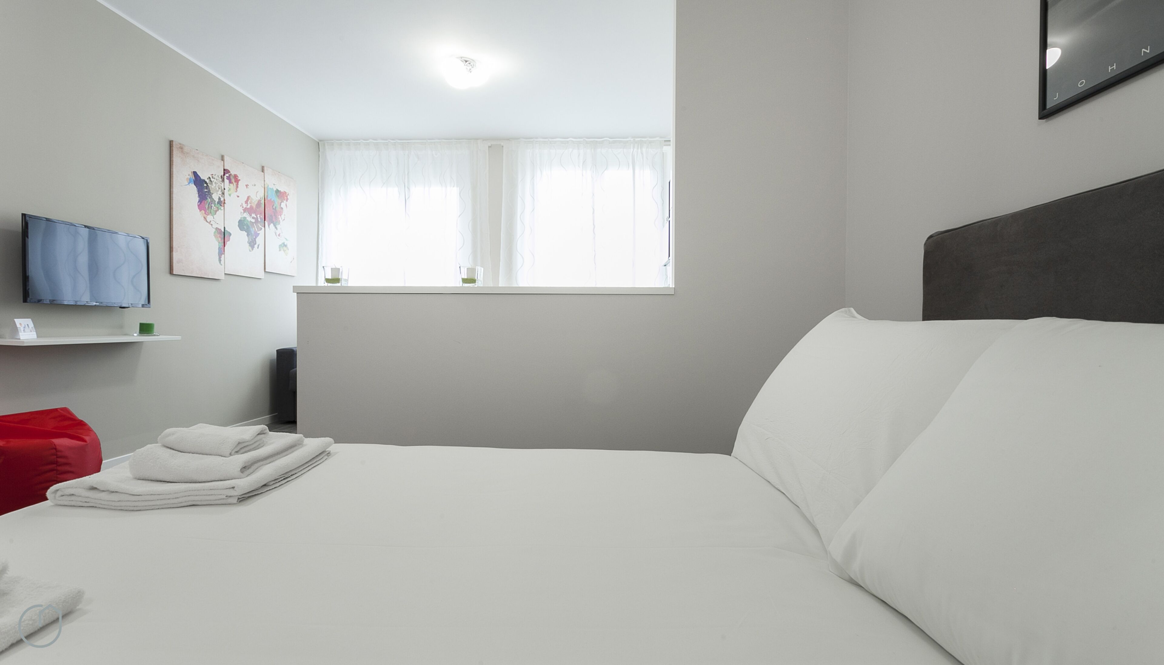 A modern hotel room with a neatly made bed, a red bean bag chair, a flat-screen TV mounted on the wall, and a window with sheer curtains.