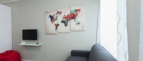 A modern living room with a grey sofa, a red bean bag, a flat-screen TV on a white shelf, and two abstract world maps on the wall.