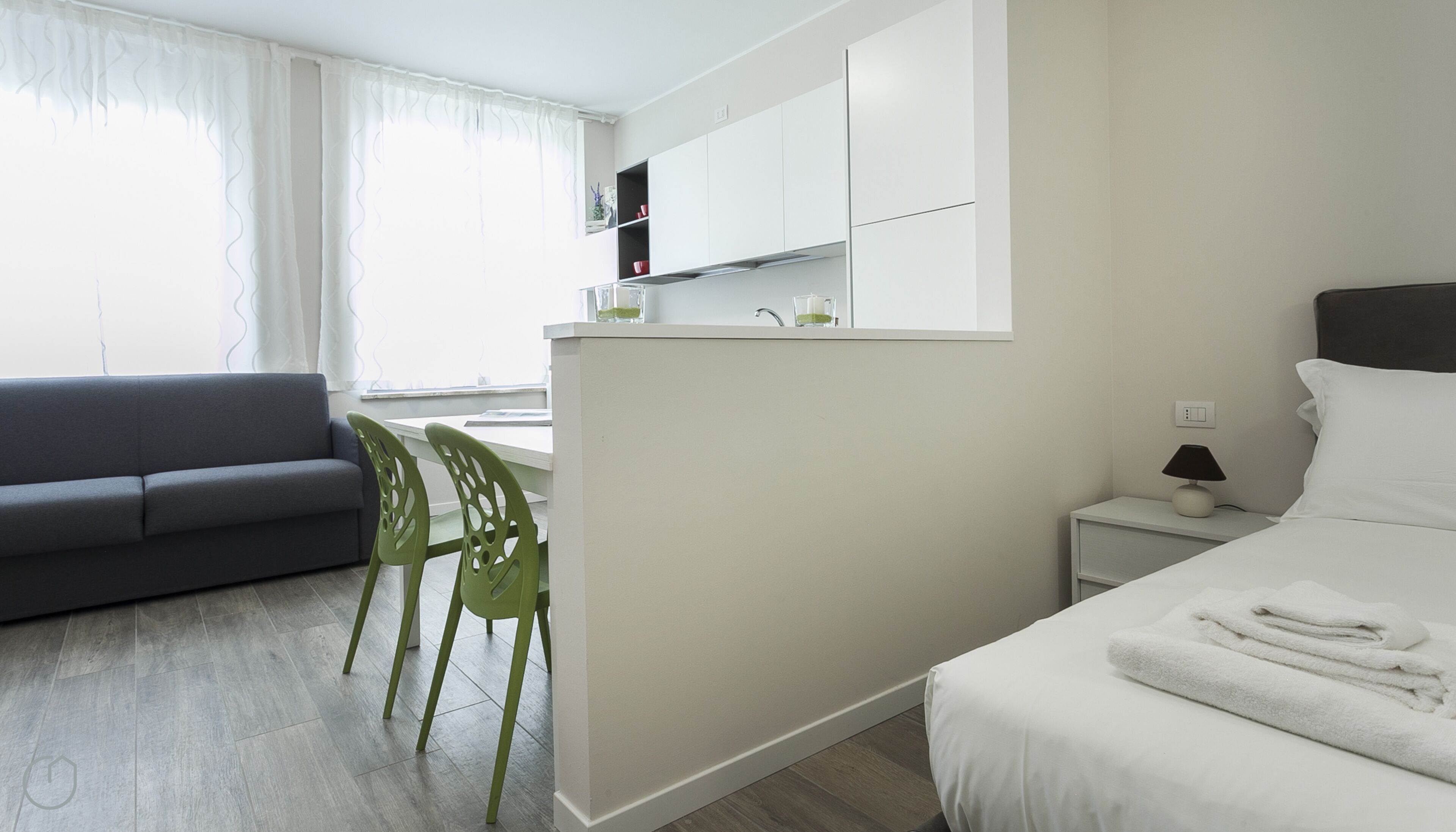 Ett modernt hotellrum med en grå soffa, en grön stol, en säng med vita sängkläder, ett litet sängbord med en lampa och ett litet pentry med vita skåp.