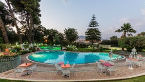 Una piscina circular con sillas y mesas blancas alrededor, rodeada de árboles y vistas a un paisaje urbano.