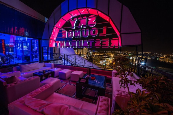 A rooftop lounge area with a neon sign reading "SNY LOUNGE" and a city view.