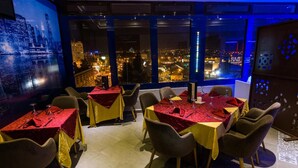 Un restaurante con mesas dispuestas para comer, amplios ventanales con vista a la ciudad y un mural de paisaje urbano en la pared.