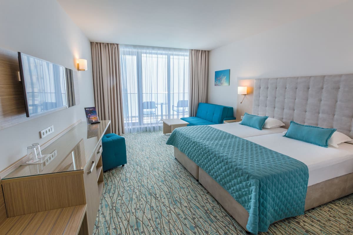 Ein Hotelzimmer mit einem großen Bett, einem Schreibtisch, einem Sessel, einem Fernseher und einem Balkon mit Vorhängen.