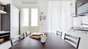 Uma área de refeições com uma mesa de madeira, cadeiras brancas, uma caneca branca e uma estante.