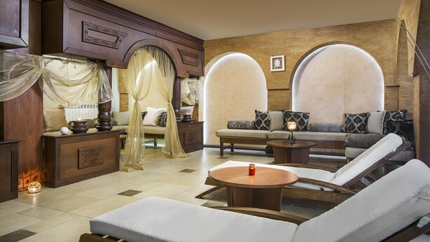 A spacious living room with a sofa set, a coffee table, and a chaise lounge. There are two large arched windows with curtains. - Royal Park Hotel & Apartments (Bansko)