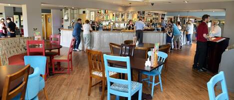 Zona de bar con mesas y sillas de madera, una barra y un grupo de personas.