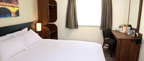A hotel room with a large bed, a desk with a chair, a mirror, a window with curtains, and a painting on the wall.