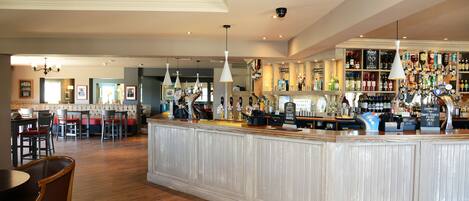 A bar area with a wooden counter, hanging pendant lights, and a variety of bottles and glasses.