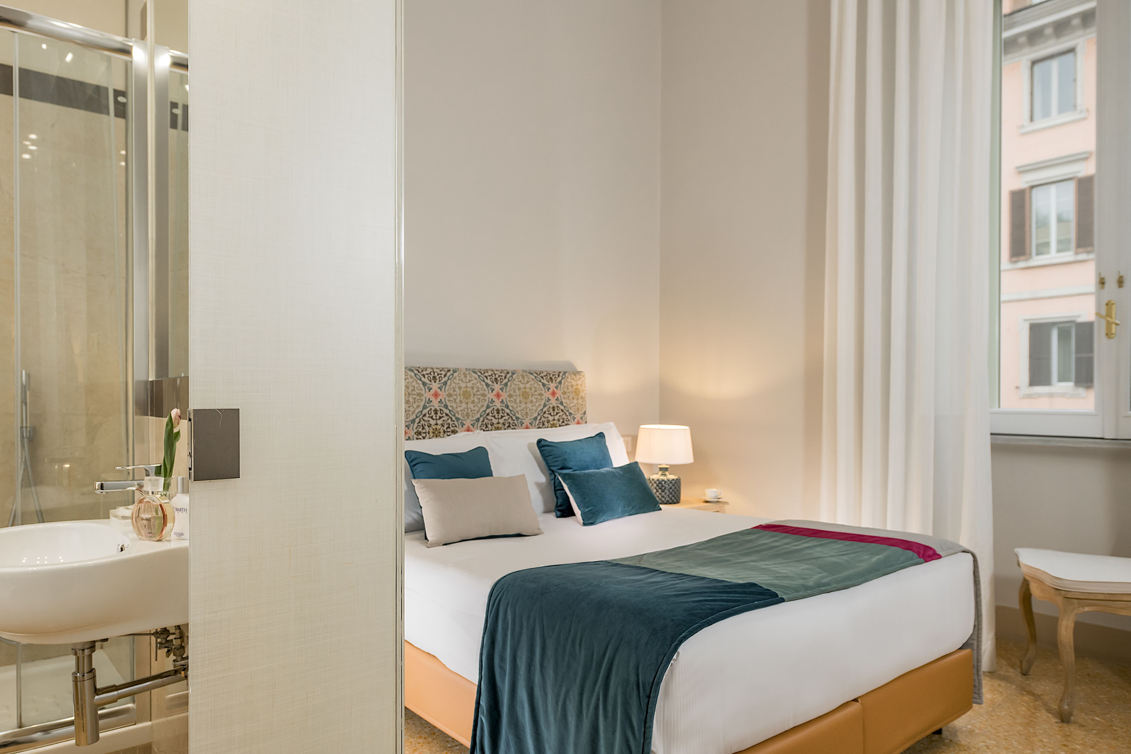 A hotel room with a bed, bedside table, lamp, and a view of the outside.